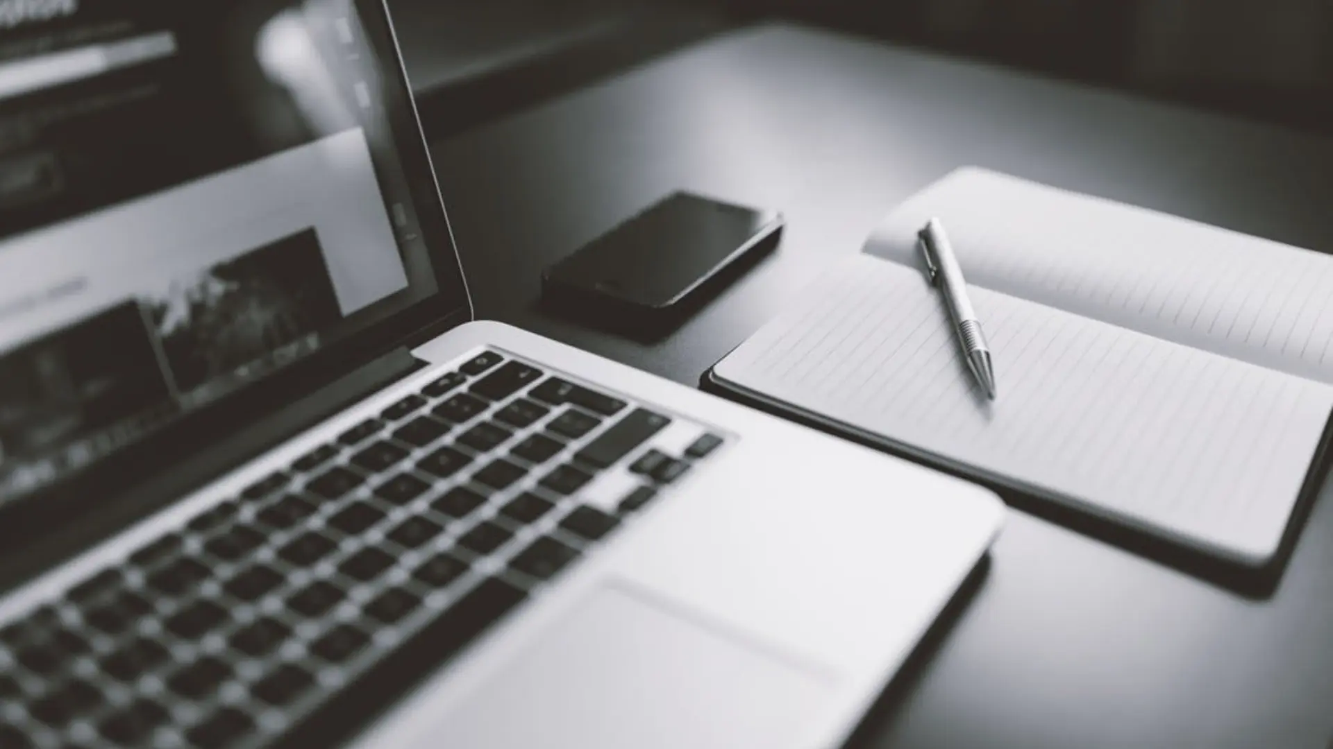 Laptop, with a phone and notebook and pen on a desk in black and white
